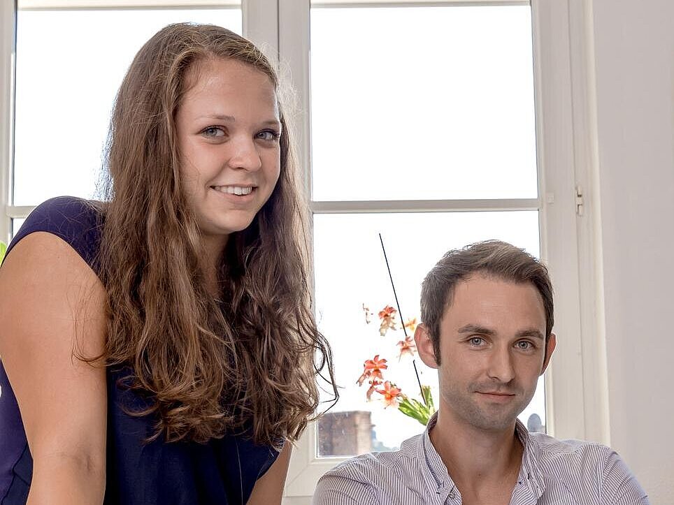 ein junger Mann sitzt am Schreibtisch, neben ihm steht eine junge Frau
